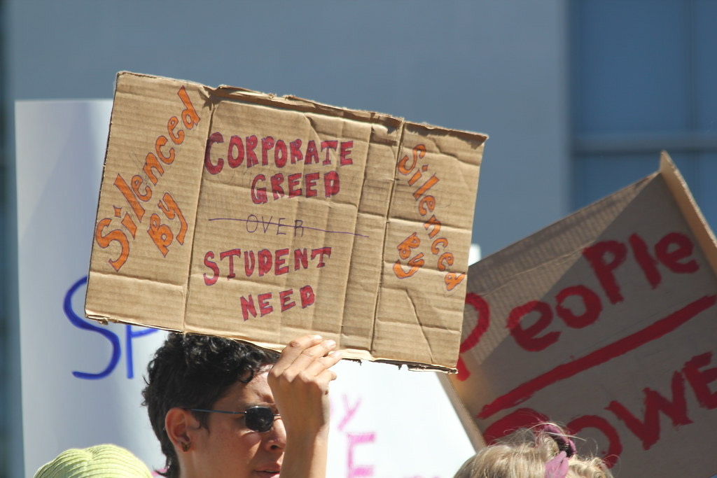 A protester at a rally holds a hand-lettered cardboard sign reading Corporate Greed Over Student Need
