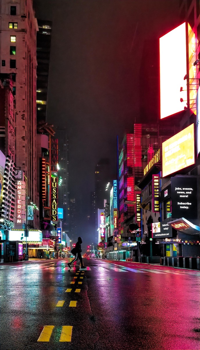 Night photograph looking down an empty 42nd Street in New York, neon signs blazing over wet tarmac, with one lone figure crossing the road.