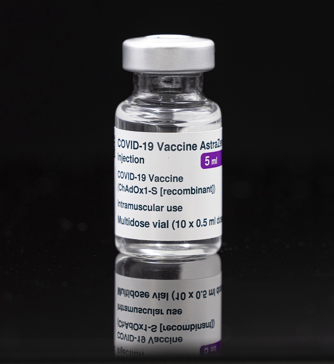 A 5 ml glass vial of COVID-19 vaccine photographed against a black background with its reflection below.